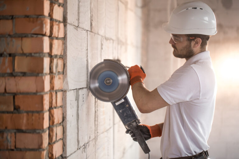Worker in a hard hat cutting through a concrete and brick wall with a grinder during the removal of a load-bearing wall.